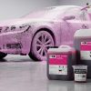 Активатор піни Foam Booster у мірній склянці на тлі каністр автохімії та автомобіля, що покритий щільною рожевою активною піною під час миття.