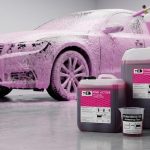 Активатор пены Foam Booster в мерном стакане на фоне канистр с автохимией и автомобиля, покрытого густой розовой активной пеной во время мойки.